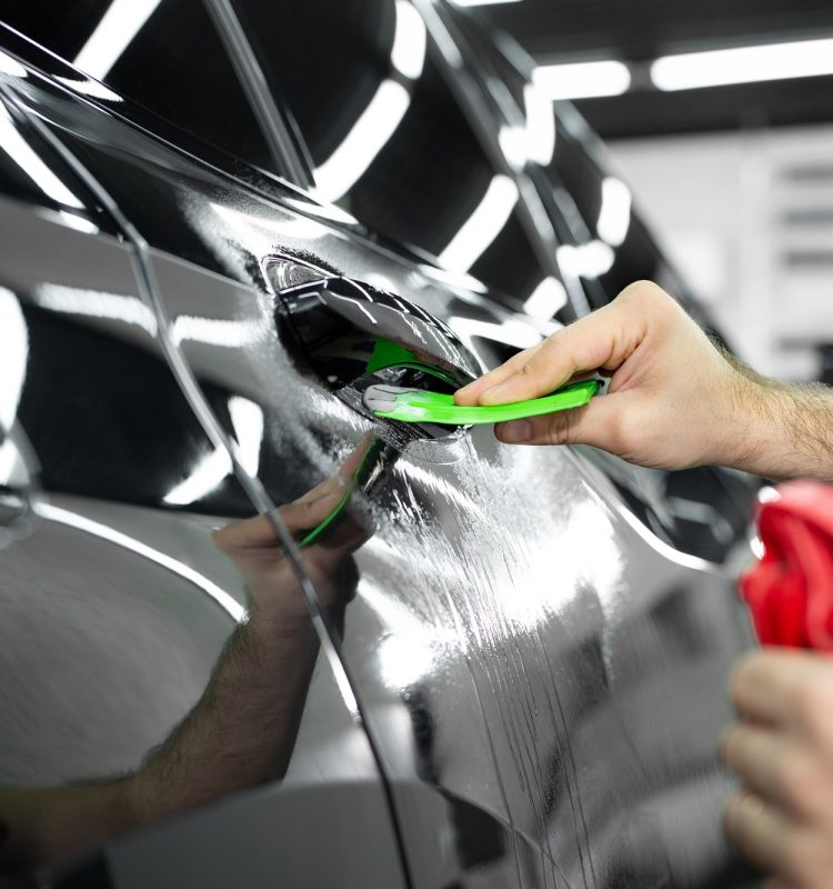 Car service worker glues anti-gravity film on the car body with a scraper in car detailing workshop